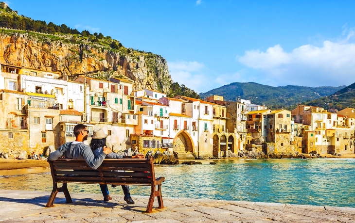 Paar sitzt auf der Bank, genießen Blick auf Strand Stadt Cefalu in Sizilien, Italien